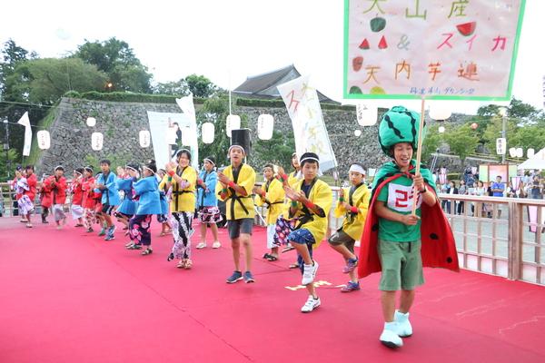 黄色、青、赤の法被を着た生徒と、スイカの帽子とマントをした男性がプラカードを持って踊っている写真