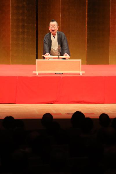 落語家の桂文珍さんが真っ赤な布に覆われた舞台の上で観客にお話ししている写真
