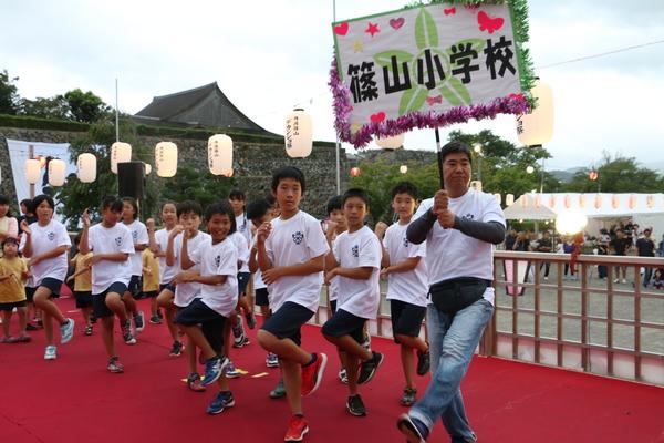 篠山小学校と書かれたプラカードを持った男性と、体操服を着た生徒が躍っている写真