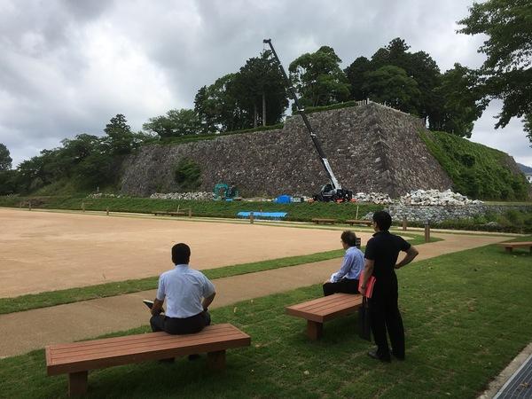 クレーン車が作業しているところを関係者の男性3名がベンチに座り見ている写真