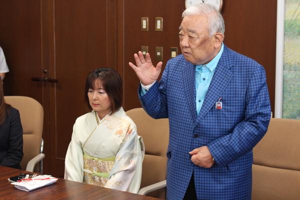 左側に着物姿の女性が座りその右側に青いチェックの上着を着た男性が立ち上がって話をしている写真