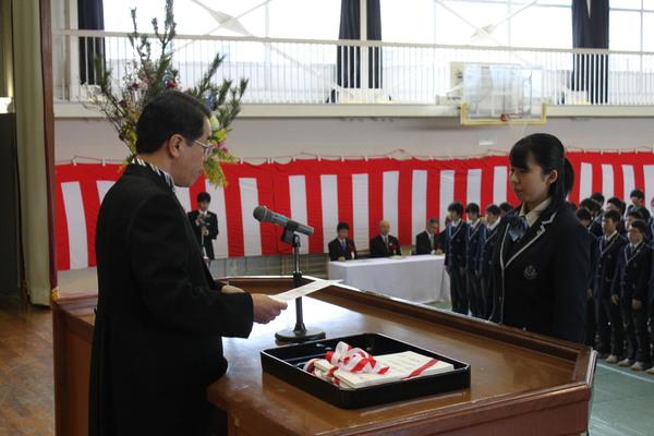 ステージ上の壇上でマイクの前で校長が女子生徒に向けて卒業証書を読み上げている写真