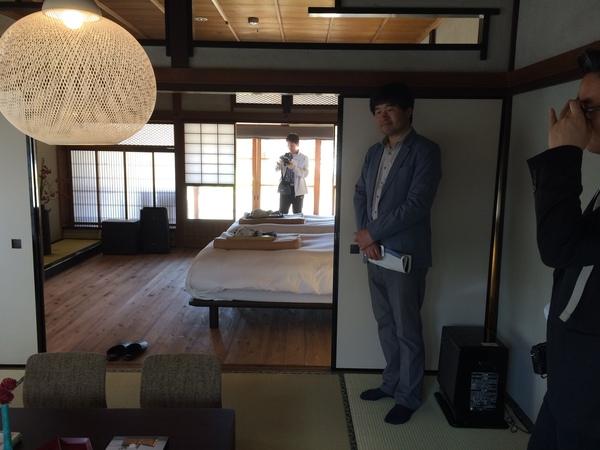 ベットが置かれた部屋の隣の、畳の部屋に立つスーツの男性の写真
