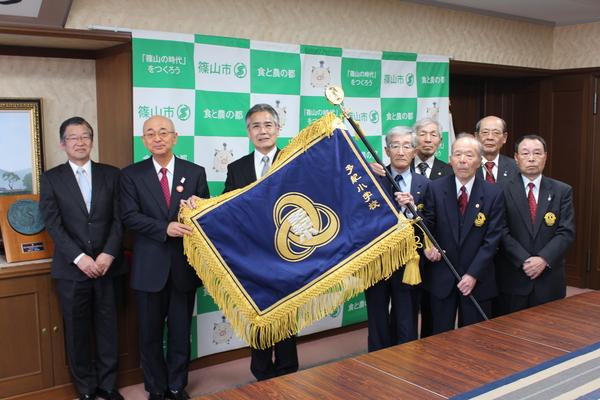 多紀ライオンズから寄贈された青色の布に金色の校章が入っている校旗を広げて見せて、市長と関係者が一緒に写っている写真