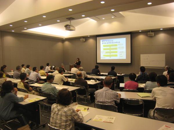 モニターの横で講師の女性が話をしていて、受講者達が椅子に座って話を聞いている写真