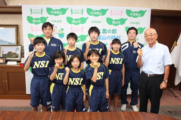 選手7名と両脇後方にコーチ2名と右端に市長が右手を曲げて拳を握るポーズを取り記念撮影をしている写真