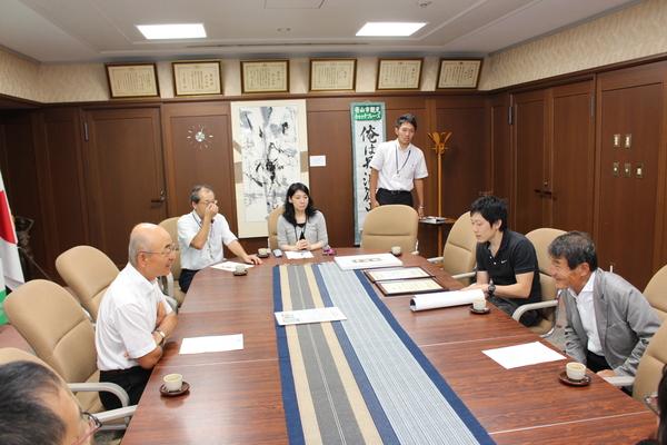 長い机に座り園田雄一さんの前に置かれている賞状をみていて、市長と話をしている写真