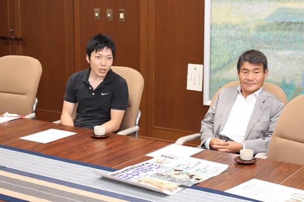 黒の半袖姿の園田雄一さんが話をして、その隣に背広姿の男性が座っている写真