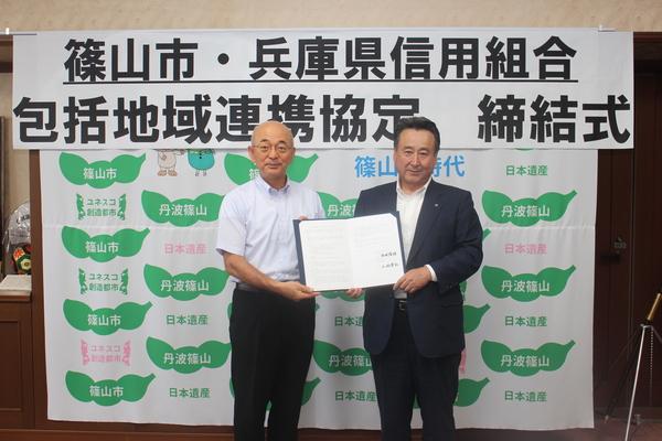 「包括地域連携協定 締結式」と書かれた幕の前で市長と土肥理事長が二人で協定書を持って並んでいる写真