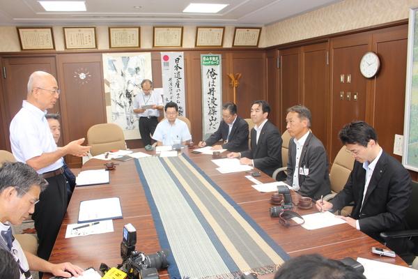 楕円形のテーブルに7名ほどのスーツを着た男性達が座り市長が立って話をしている写真