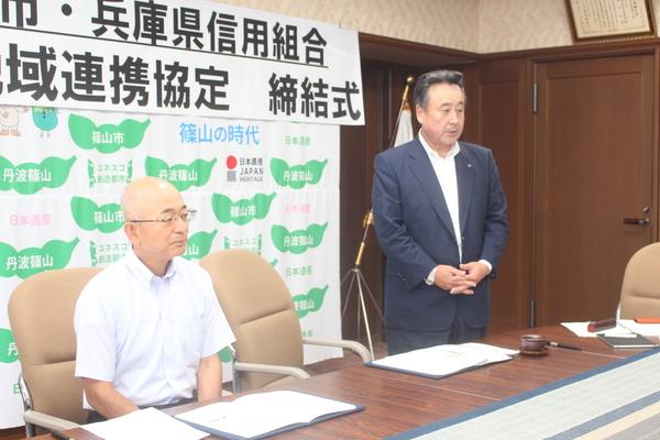 市長が左側に座っていて、右側で土肥 貴弘理事長が立って話をしている写真