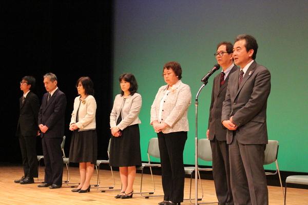 退職者7名が立っていて、男性がスタンドマイクで挨拶をしている写真