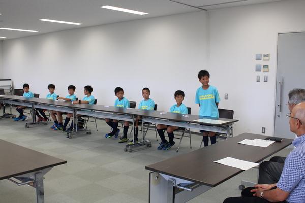 水色のユニフォームを着た男の子達が、着席しているなか、1人の男の子が立っている写真