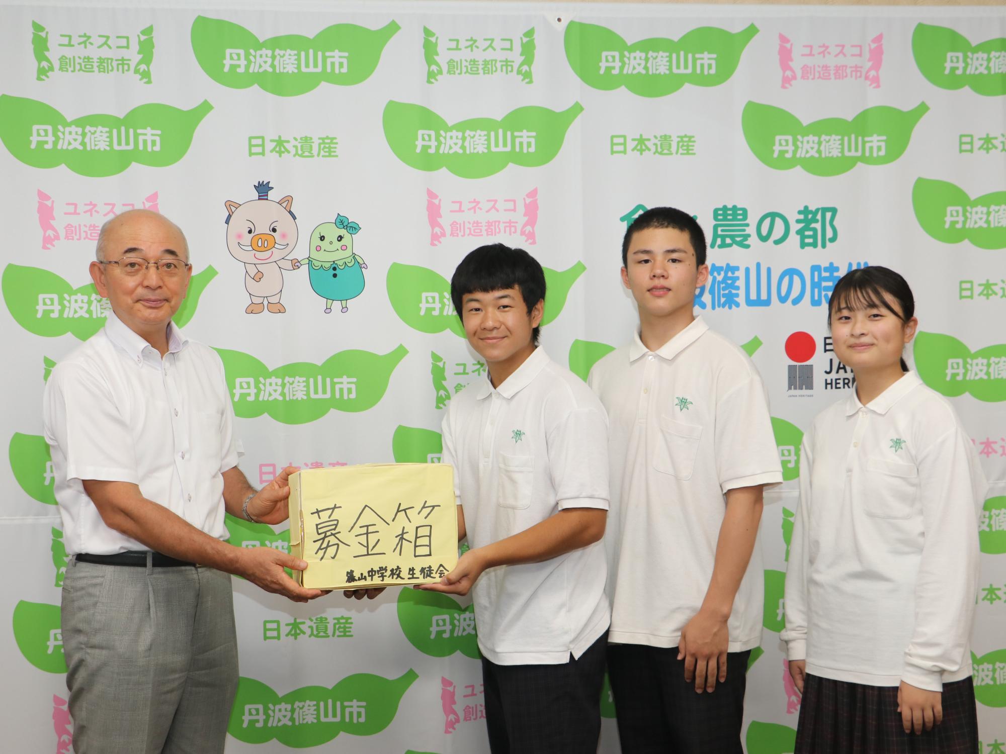 制服を着た男子中学生が、募金箱と書かれた黄色の箱を市長に手渡している。隣には男子中学生と女性中学生が立っている。