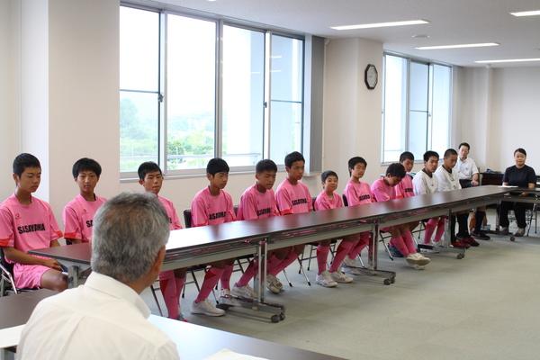 着席しているピンクと白色のユニフォームを着た篠山、丹南中学校男子ホッケー部の男子学生と、男性の写真