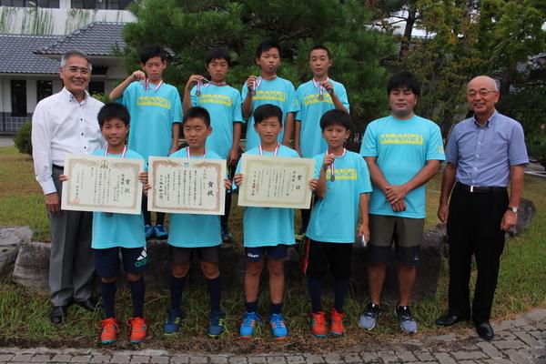 青いユニフォームを着た少年達が市長と記念撮影、3名の男の子が賞状を手に他はメダルを手に持っている写真