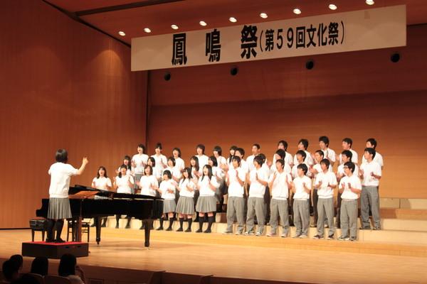 ホールで生徒たちが指揮者を見ながら手拍子をして歌っている写真