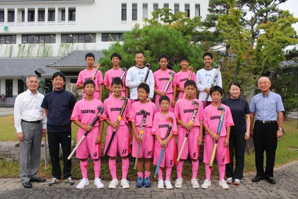 ピンクのユニフォームを着た子供達がホッケーを手に、市長と笑顔で記念撮影写真