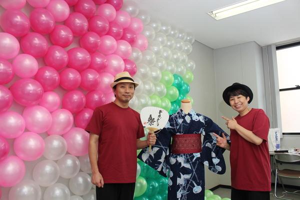ピンクと白の風船が飾られた壁の前で浴衣を着たマネキンの横に立つ2名の男女の写真