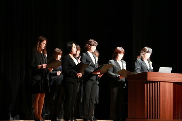 上下黒のスーツを着て手に資料も持っている複数の女性たちが壇上に立ち、そのうちの1名が話をしている写真