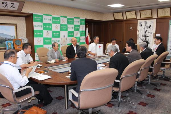 日本国旗や、掛け軸が掛けられている応接室で市長や10名ほどの人達が座って話をしている写真