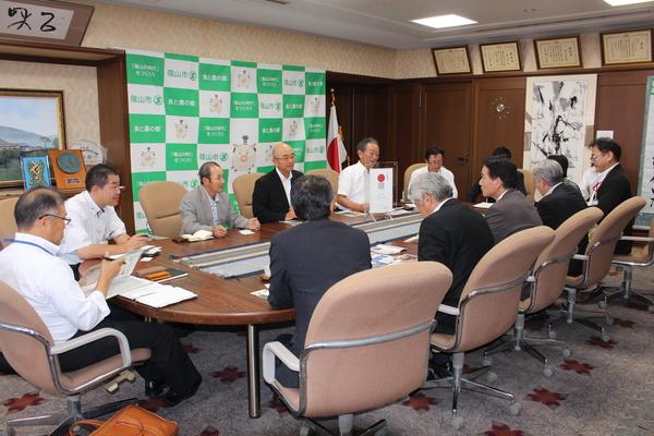 日本国旗や、掛け軸が掛けられている応接室で市長や10名ほどの人達が座って話をしている写真