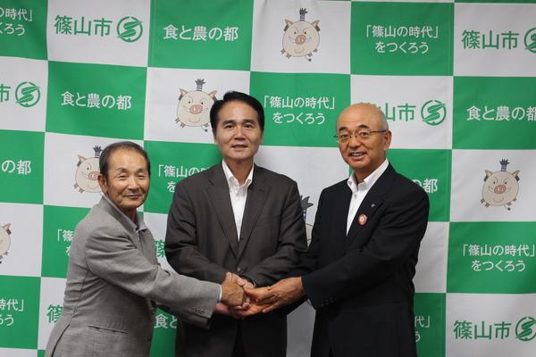 右はじには市長、その横に2人のスーツを着た男性が並んでいて、3人が中央で握手をしている写真