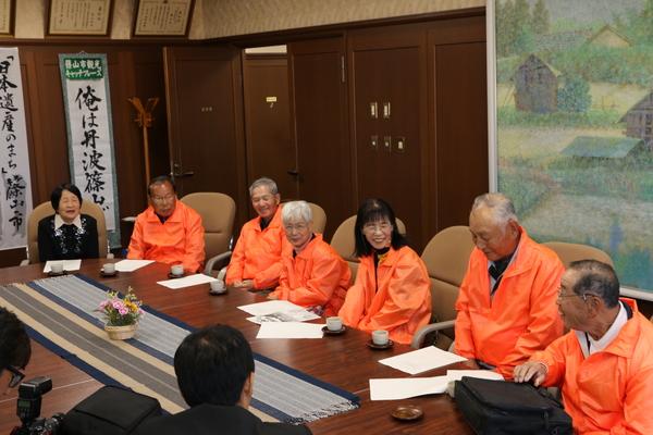 オレンジ色のジャンパーを着た篠山市身体障害者福祉協議会のメンバーが椅子に座っており、会長の山本 清さんが話をしているのを、笑顔でみている様子の写真
