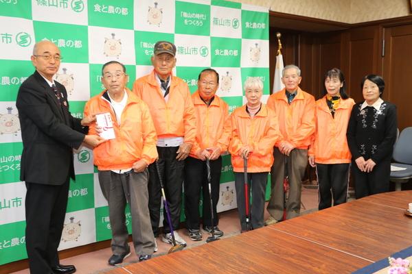 篠山市身体障害者福祉協議会の皆さんがオレンジ色のジャンパーを着ており、会長の山本 清さんが市長より奨励金を手渡しでもらっている様子の写真