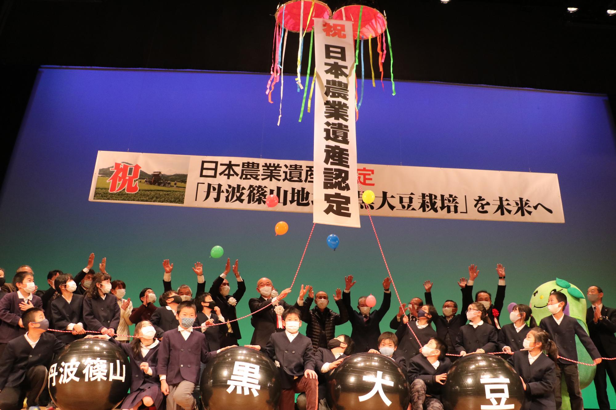 舞台上で、日本農業遺産認定のくす玉を関係者のみなさんでひもをひっぱって割っている。