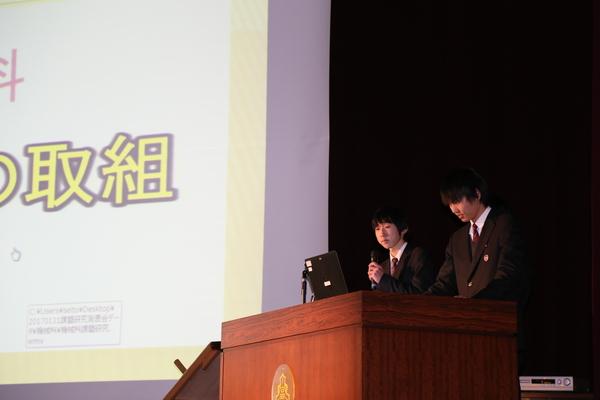 舞台の上のプロジェクタースライドの横で2人の高校生がパソコンを見ながらマイクを持っている写真