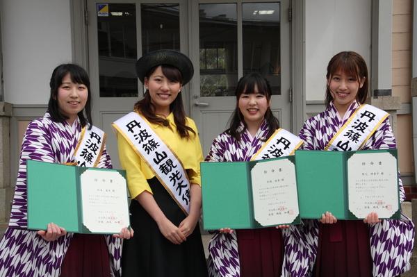 袴を着る丹波篠山観光大使が、任命書を持ち、制服と帽子をかぶり、観光大使のタスキをつけた女性が笑顔で並ぶ写真