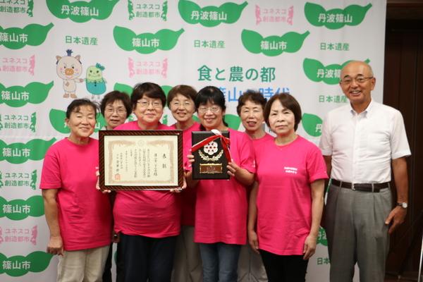 受賞の賞状が入っている額を持った女性と、表彰盾を持っている女性の周りに篠山市いずみ会の他5名が立ち、市長と一緒に写っている写真