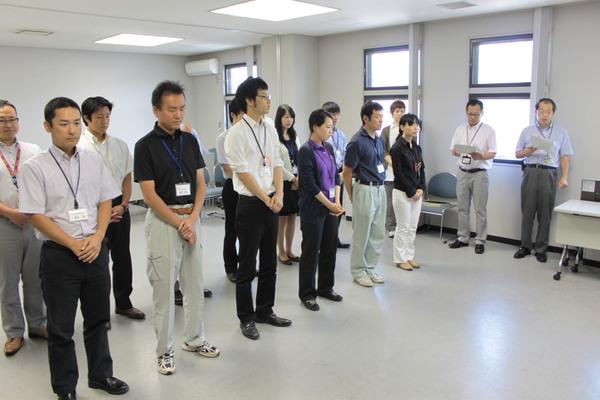 窓際で、男性2名が書類を手に持ち1人が話をしているのを2列に並んでいる職員の方達が立って聞いている写真