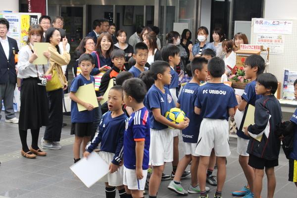 市役所のロビーにサッカーのユニフォームを着た少年たちが手にサッカーボールや色紙を持って立っている写真