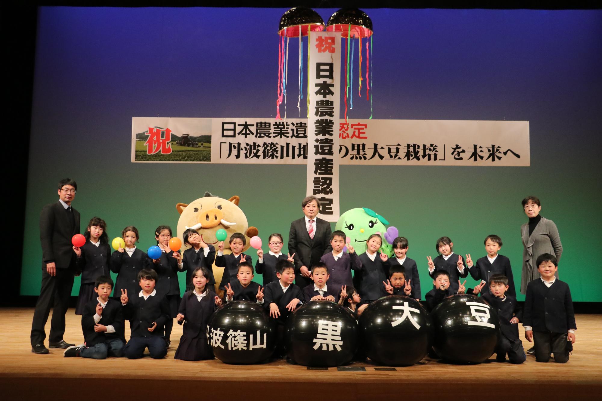 日本農業遺産認定を祝うくす玉と、小学校のこどもたち、まるいのとまめりんが舞台で記念撮影している。