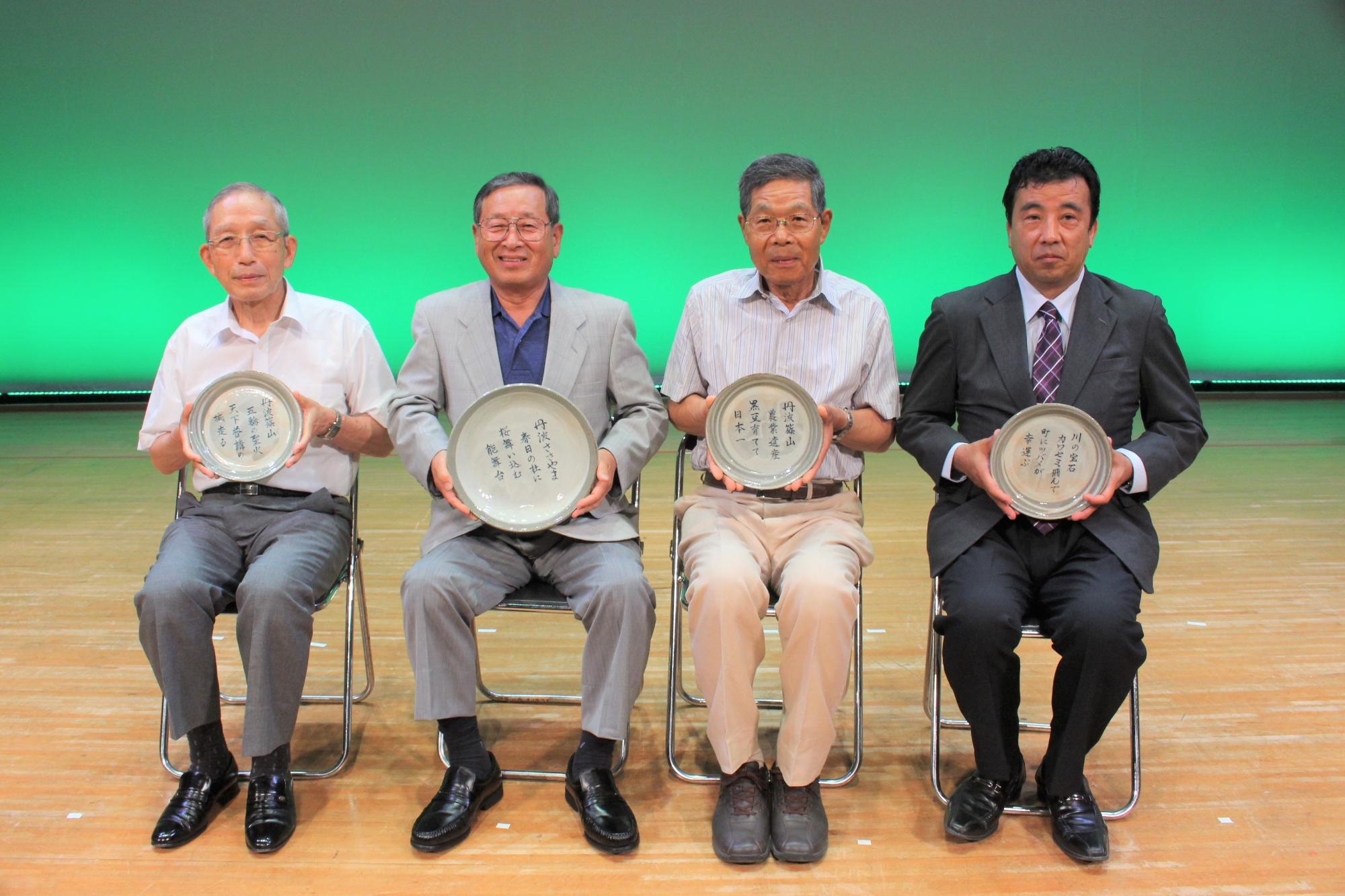 受賞された歌詞が書かれた丹波焼の器を4人の受賞者が椅子に座って持っている