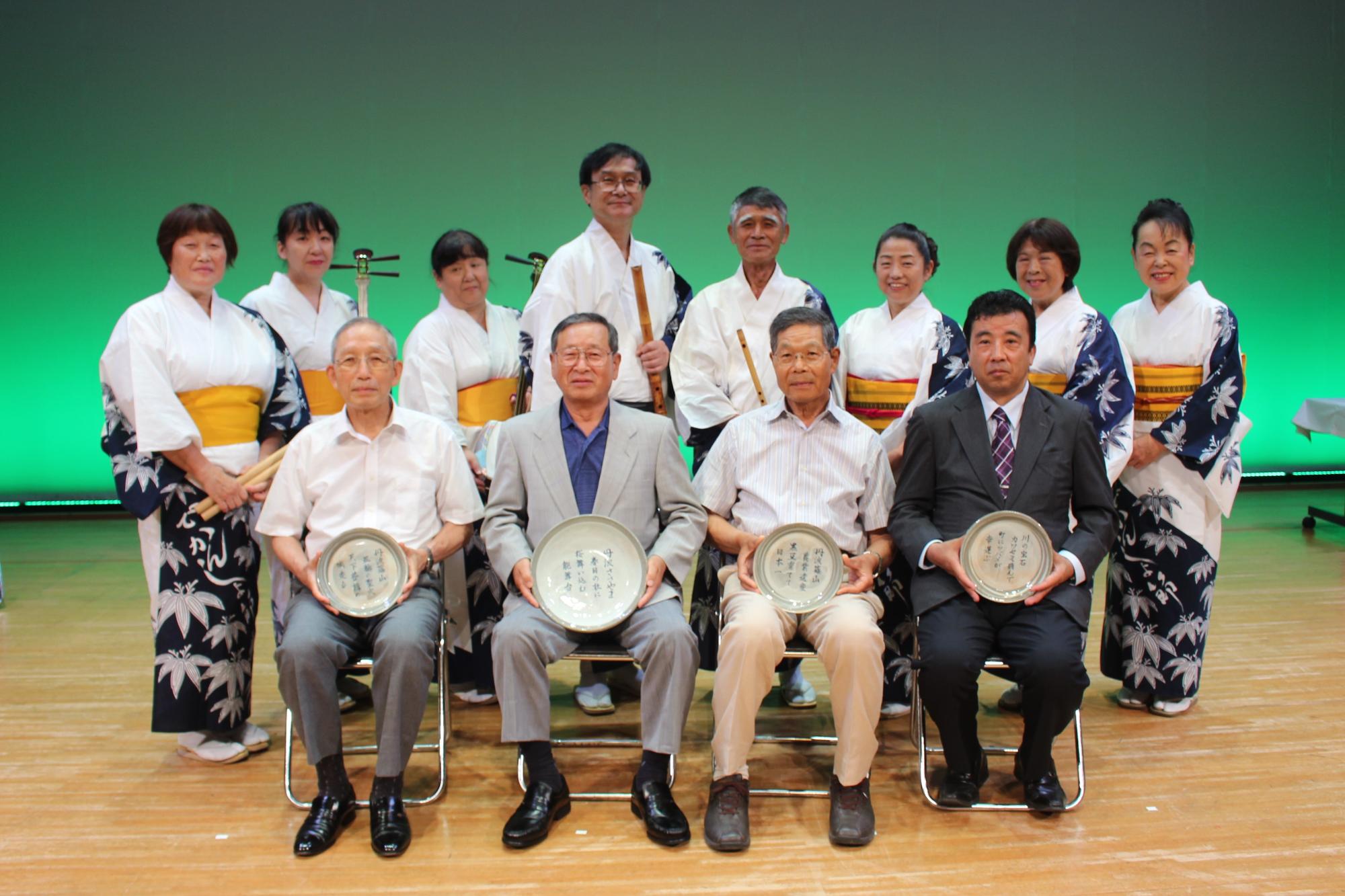 デカンショ節を演奏しうたわれる地方が後ろに立ち、前方に受賞者4名が椅子に座っている
