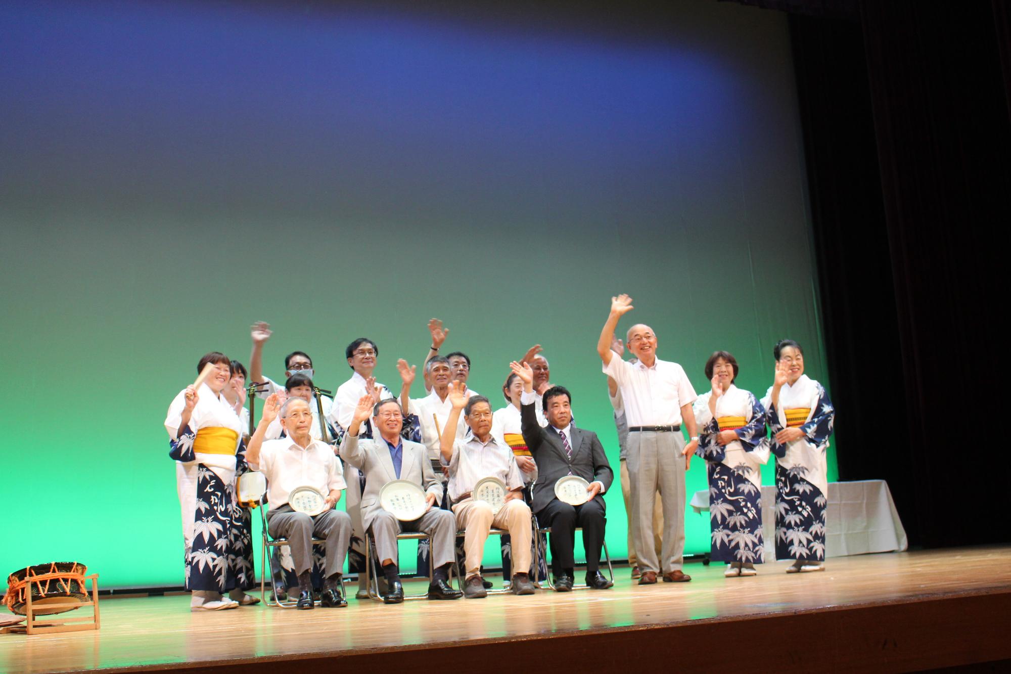 受賞者が椅子に座り、市長の地方がその周りにたち、皆で手を振っている