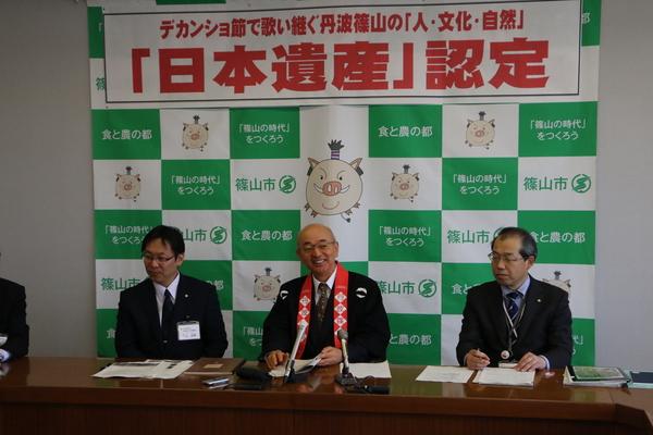 「日本遺産」認定と書かれている横断幕の前で、市長が法被を着てマイクの置かれている机の前に笑顔で座っている様子の写真