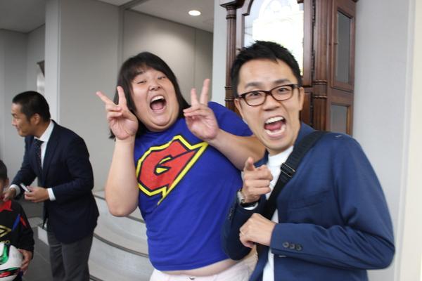 青いGのマークの入ったティーシャツを着ているガリガリガリクソンさんが両手でピースサインをして、青いジャケットを着た女と男の市川さんが右手で指さして写っている写真