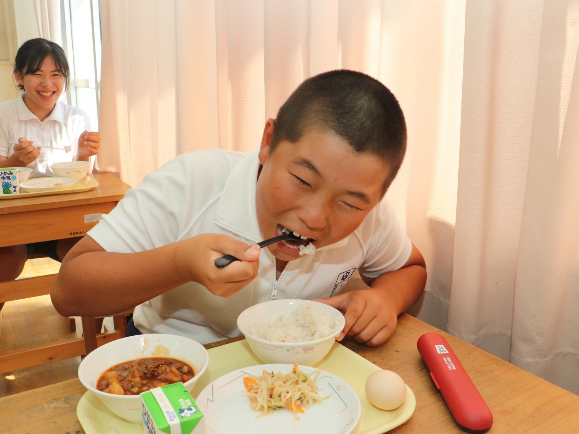 男の子が勢いよく給食のご飯を食べている。後ろの座っている女の子がその姿を見て笑っている。