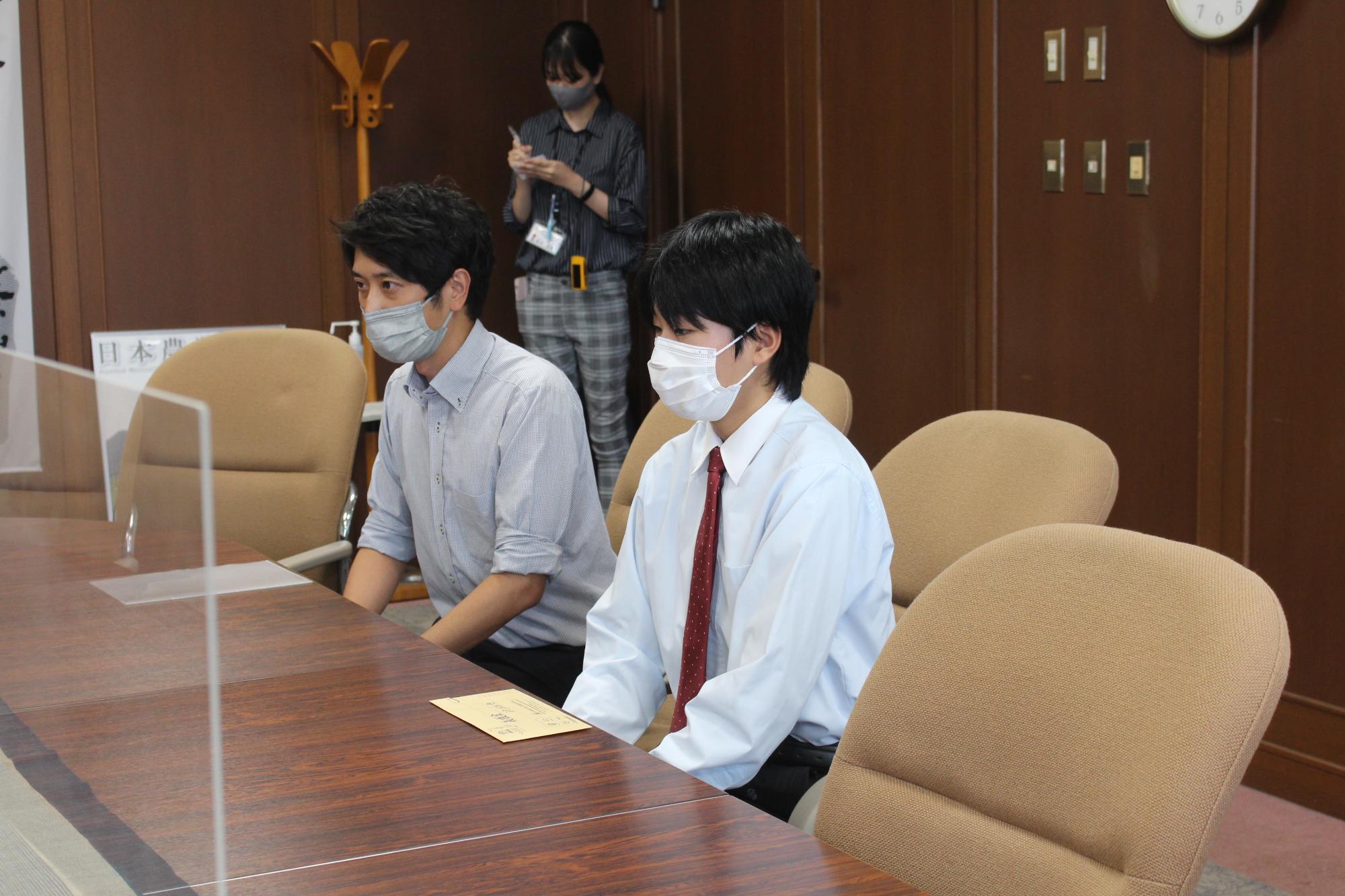 椅子に二人の男性が座っていて、テーブルには義援金が入った封筒が置いてある。