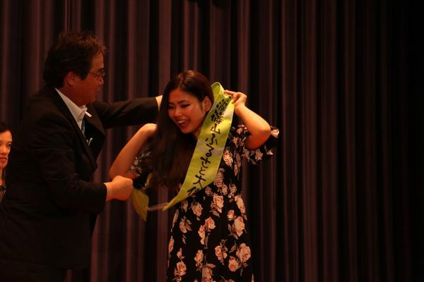 遠山 恵さんが、丹波篠山ふるさと大使のタスキをスーツの男性からもらっている写真