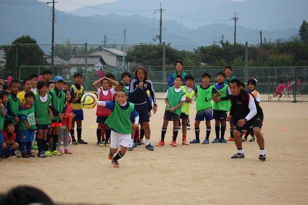 緑のゼッケンの少年がボールを追いかけていて、その後ろで北澤選手がマイクを持って話をしている写真