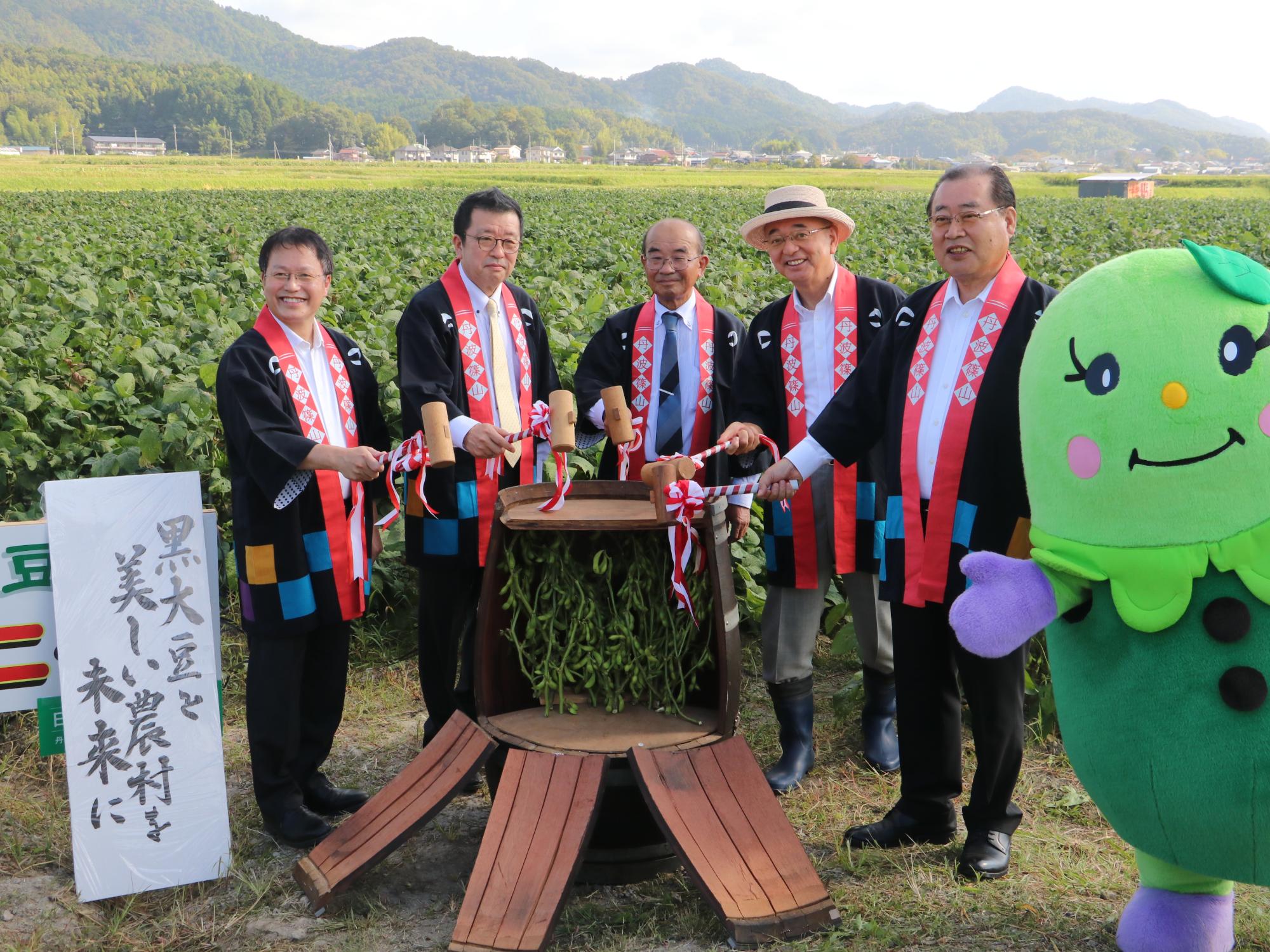 黒豆畑の前で、ハッピを着た6人の男性が枝豆の樽をわっている。横にはまめりんが立っている。