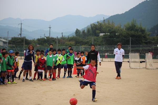 赤いゼッケンの少年がボールを蹴ろうと足をあげていて、その後ろに北澤選手や少年達が横一列に並んで立って見ている写真