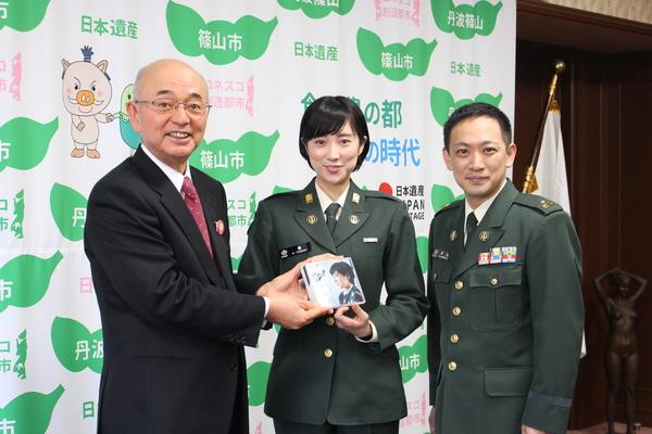 市長と緑色の自衛隊員の礼服を着た鶫 真衣さんが、鶫さんのCDを持ち自衛隊員の礼服を着た柴田隊長と3人で写っている写真