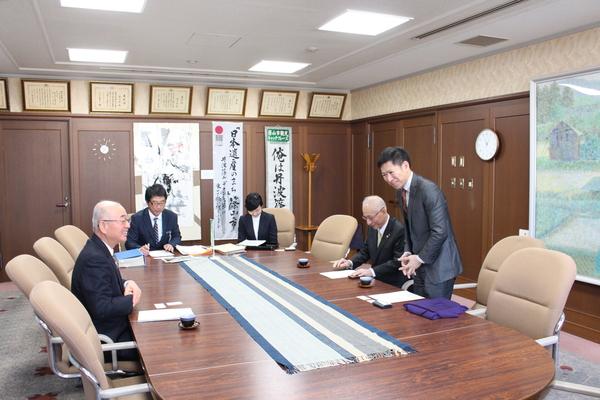 楕円形のテーブルの椅子に市長が座りその周りに4名の人達がいて、市長の向かいの席の人が立ち上がって話をしている写真