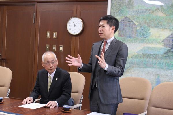 テーブルの左側にはスーツを着た男性が座っていて、右側にはスーツを着た男性が立って話をしている写真